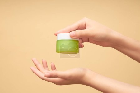 Photo for Female hand holding a green glass jar about to place on the palm of her other hand on beige background. Glass without label, mockup for cosmetic product. Front view, concept of skincare. - Royalty Free Image