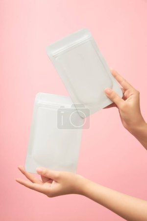 Photo for Female hands is showing two white masks packaging with no label on pastel pink background. Mockup for cosmetic product for face, cleansing, moisturizing and whitening. - Royalty Free Image