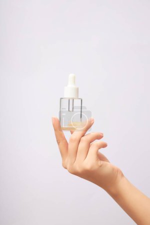 Glass serum bottle with dropper cap in woman's hand. Labelless clear glass bottle containing colorless essence on light background. Mockup for cosmetic product, front view.