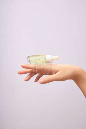 Front view of an unlabeled glass bottle containing light yellow essence, placed on the back of a woman's hand on light background. Mockup for package design, concept of skin care