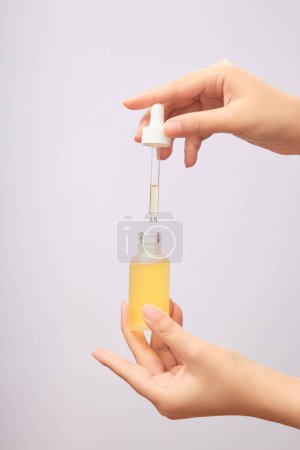 Glass bottle of serum with dropper cap, containing yellow essence in women's hands on light background. Advertising photo for cosmetic product, skincare concept.