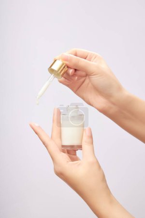 Female hands holding a white serum bottle without label, dripping of serum on index finger on light background. Skincare, cosmetic beauty product for face.