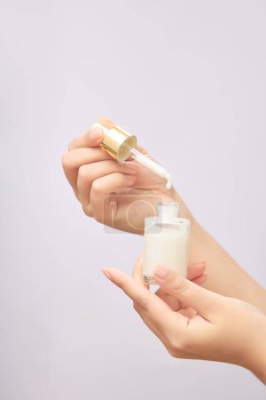 Beautiful female hands holding a bottle of serum without a label, drop by drop on the serum bottle on a light background. Cosmetic beauty product branding mockup.