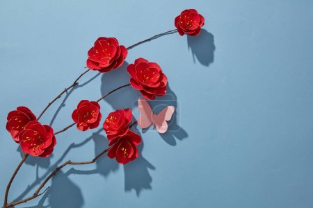 Photo for Frame with red paper flower branch, pink butterfly and their shadow on blue background. Spring flowers concept. Space for text. Top view. - Royalty Free Image