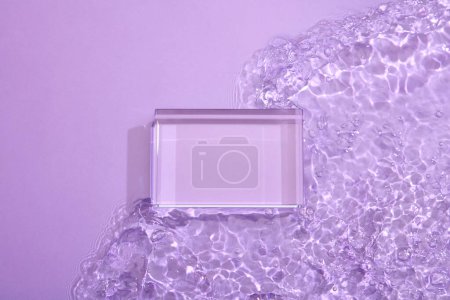 Rectangular transparent empty podium set on purple transparent clear water surface texture with ripples, splashes and bubbles. Minimal art background with copy space for cosmetics presentation. Top view, flat lay.