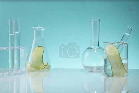Foto de Los utensilios de vidrio de laboratorio que contienen rodajas de pepino están decorados con un fondo azul. Espacio en blanco para mostrar el producto de belleza natural del extracto de pepino (Cucumis sativus) - Imagen libre de derechos