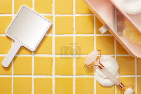 Photo for A hand mirror arranged with cotton pads, rose quartz facial massage roller and a mini plastic box with drawers. Blank space on mosaic tiles background for beauty product promotion - Royalty Free Image