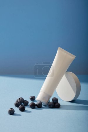 Photo for A cosmetic tube leaning on a round pedestal decorated with several blueberries over blue background. Blueberry (Vaccinium Corymbosum) extract is rich in Omega 9, an excellent carrier - Royalty Free Image