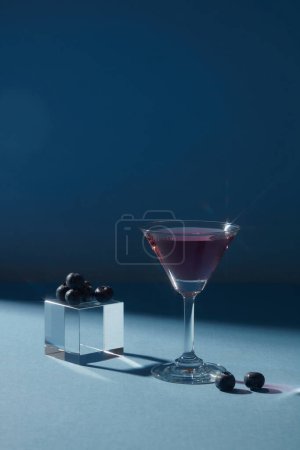 Photo for Transparent cube with blueberries placed on, decorated with a cocktail glass of purple liquid over a dark background. Promoting product extracted from Blueberry (Vaccinium Corymbosum) - Royalty Free Image
