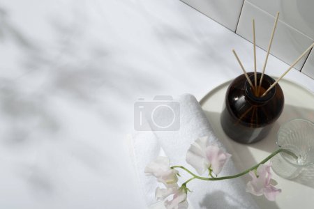 A reed diffusers, a towel and glass transparent vase with flower branch placed on a dish with round shape. Copy space with natural shadow, empty space for product presentation