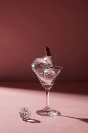 Photo for A transparent cocktail glass containing diamond sphere and an elegant red lipstick in silver package. Dark background for lady night concept - Royalty Free Image