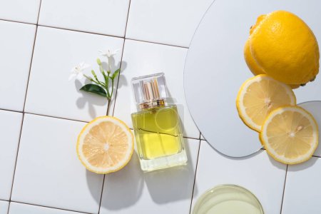 Against a white mosaic tiles background, a perfume transparent bottle displayed with flower branch, mirror and a petri dish of yellow liquid. Lemon (Citrus limon) essential oil creates a feeling of comfort