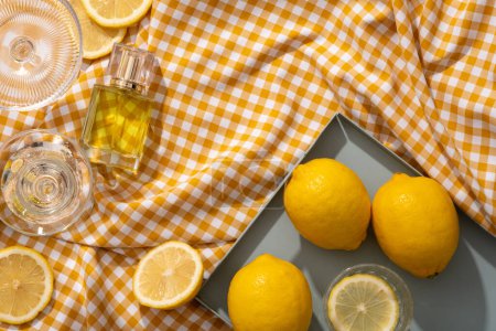 Empty label perfume glass bottle arranged with some glasses and a tray of Lemons. Vacant space on white and orange checkered fabric for organic beauty product promotion of Lemon (Citrus limon) extract