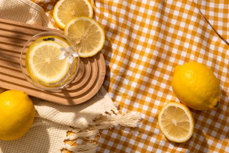 Téléchargez les photos : Un verre contenant une tranche de citron une fleur blanche placée sur un plateau en bois et un foulard en laine. Espace vide pour la publicité de produits cosmétiques bio d'extrait de citron (limon d'agrumes) - en image libre de droit