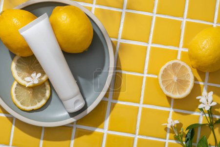 Foto de Etiqueta vacía tubo blanco, rodajas de limón y pequeña flor poner en un plato redondo. Limón (Citrus limon) puede reducir la inflamación y el exceso de producción de sebo. Mosaico amarillo azulejos fondo - Imagen libre de derechos