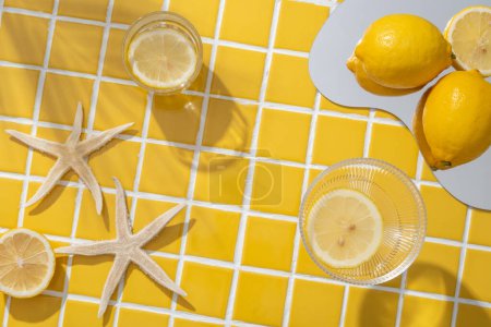 Foto de Limón colocado en un espejo arreglado con estrellas de mar y dos vasos que contienen rodajas de limón. Espacio vacío en el fondo de azulejos de mosaico amarillo para mostrar producto cosmético natural - Imagen libre de derechos