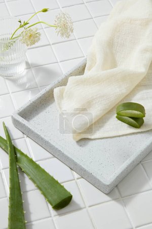 Ein Tablett mit Handtuch und Aloe-Vera-Scheiben, darauf ein transparentes Glas mit weißen Blüten. Leere Flächen für die Produktpräsentation. Konzept natürlicher Schönheitsprodukte