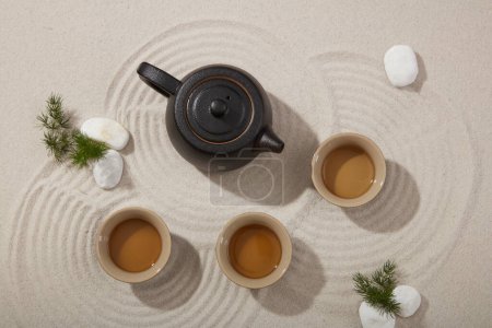 A set of tea with an earthen teapot and three tea cups, arranged with white gravels and green leaves. Natural beach sand background. Top view