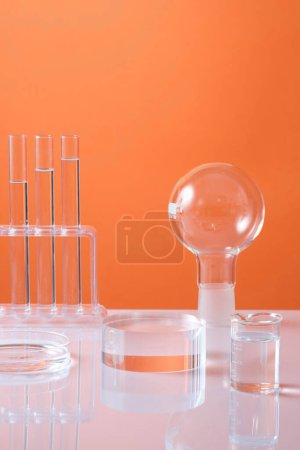 Foto de Un podio transparente de vidrio en forma de cilindro colocado en la mesa con un vaso de precipitados, una placa de Petri y otros artículos de vidrio de laboratorio. Fondo naranja - Imagen libre de derechos