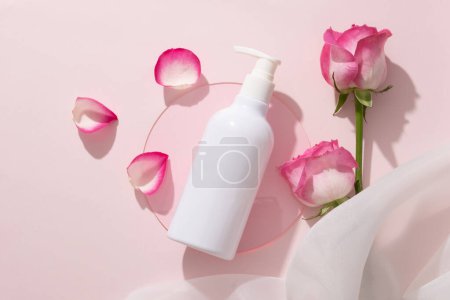 Téléchargez les photos : Vue de dessus d'une bouteille de pompe cosmétique blanche arrangée avec des roses, une feuille acrylique en forme ronde et un tissu en mousseline de soie. Maquillage de soins de la peau cosmétique de beauté faciale - en image libre de droit