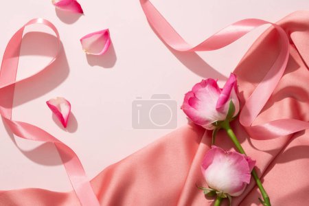 Roses are placed on an elegant fabric in pink color with pink ribbons and Rose petals. Organic product extracted from Rose (Rosa) can be displayed in the empty space