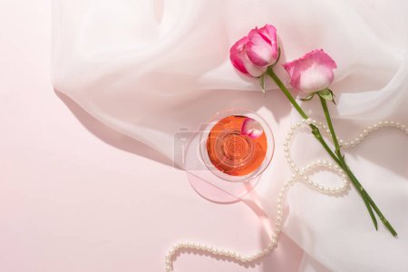 Cocktail glass filled with liquid extracted from Rose (Rosa) and a pearl necklace displayed on chiffon fabric. Blank space can be used to promote natural cosmetic product