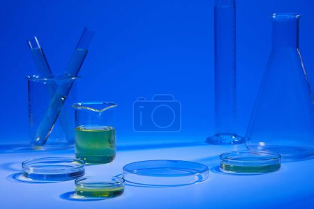 Beaker and petri dish of yellow liquid displayed with other laboratory glassware on a blue background. Vacant space on podium