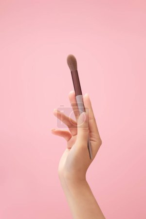 Téléchargez les photos : Un pinceau mélangeur est maintenu par un modèle de main femelle sur fond rose clair. Publicité de produits de maquillage - en image libre de droit