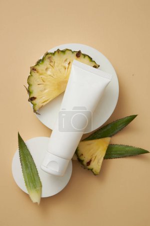 A white tube placed on round shaped podiums, displayed with pineapple leaves and slices. Empty label to promote beauty product extracted from pineapple (Ananas comosus)