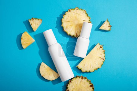 Vor blauem Hintergrund sind einige Ananasscheiben und weiße leere Etikettenflaschen zu sehen. Produktpräsentation. Ananas (Ananas comosus) verlangsamt den Alterungsprozess.