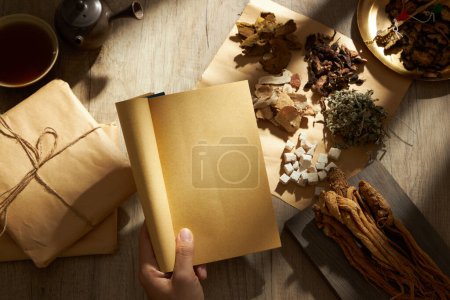 A hand model holding ancient Chinese medicine books with many types of herb, medicine packs, a bowl of medicine and earthen pot displayed behind. Herbal medicine is safe and easy to use