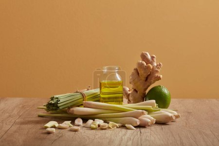 Photo for A glass transparent jar of essential oil extracted from lemongrass, ginger and lemon is displayed between bunch of lemongrass, a lemon and a ginger on wooden table - Royalty Free Image