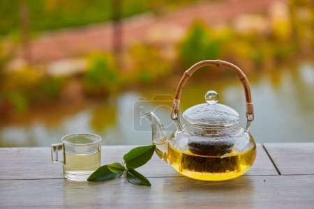 A teapot with green tea inside, a tea cup and small green tea leaves placed on wooden table, blurred background