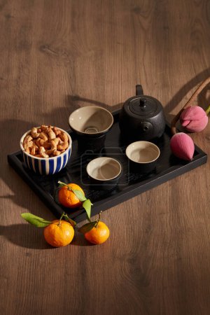 Foto de Un juego de té negro con nueces de anacardo, naranjas de mandarina y flores rosadas sobre fondo de madera. Contenido de vacaciones - Imagen libre de derechos
