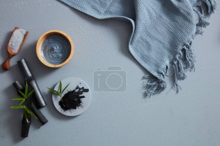 Photo for A bird's-eye view of charcoal powder, facial mask and towel on gray background. Text can be added in the blank space - Royalty Free Image