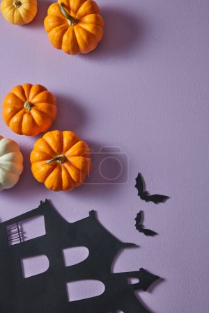A background of purple with white and orange pumpkins. Some black bats and paper house next to it. Halloween concept