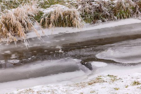 Ein völlig zugefrorener Bach mit Eis und Schnee im Winter in einer ländlichen Landschaft mit Gras am Ufer, Deutschland