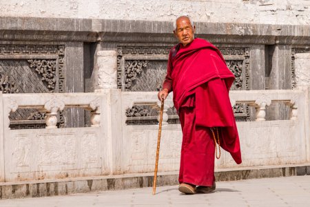 Photo for Tibetan monk in red robe with stick walks along a decorated wall of Kumbum Champa Ling Monastery, Xining, China - Royalty Free Image