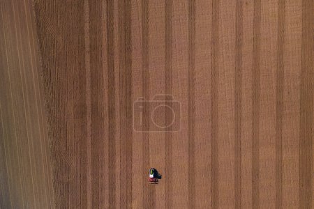 Photo for A small lonely tractor with plow on a large brown field with plowed earth seen directly from above - Royalty Free Image