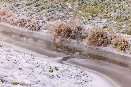 Stream frozen with ice and snow on a slope with grass in winter