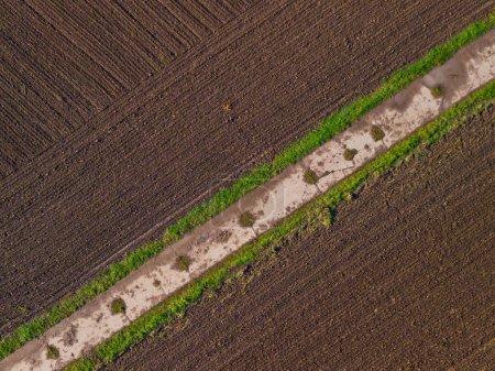 Gepflügtes Feld mit fruchtbarem Boden, das ein diagonales Muster mit einem Feldweg bildet, Luftaufnahme