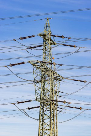 A high-voltage pylon with insulators, traverses and conductor cables as well as overhead electric lines in front of a blue sky, portrait photograph