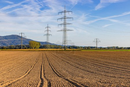 Ein landwirtschaftliches Feld mit Reifenspuren und Strommasten im Hintergrund unter blauem Himmel, Deutschland