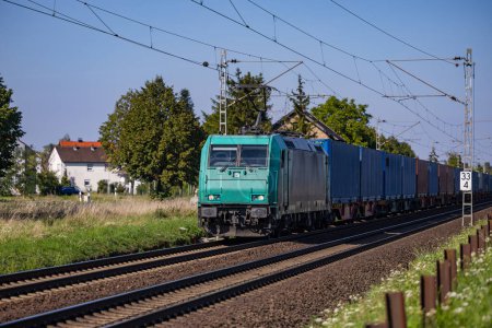 Un train de marchandises turquoise sur une ligne de chemin de fer électrique traversant une zone rurale près d'un village pour le transport public et le trafic régional de passagers