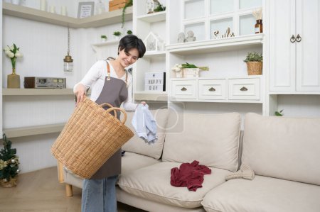 Téléchargez les photos : Une jeune femme de ménage asiatique prend des vêtements sales désordonnés dans le panier pour nettoyer à la maison, concept de style de vie bien-être - en image libre de droit