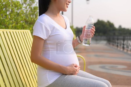 Téléchargez les photos : Belle femme enceinte buvant une bouteille d'eau pure dans le parc, concept de mode de vie de grossesse sain et actif. - en image libre de droit