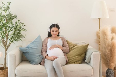 Téléchargez les photos : Joyeux femme enceinte avec écouteurs écoutant de la musique mozart et couché sur le canapé, concept de grossesse - en image libre de droit