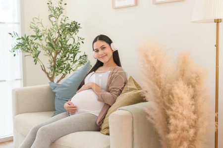 Téléchargez les photos : Joyeux femme enceinte avec écouteurs écoutant de la musique mozart et couché sur le canapé, concept de grossesse - en image libre de droit