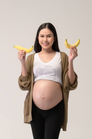 Téléchargez les photos : Portrait de Belle femme enceinte tenant banane sur fond blanc studio, concept santé et maternité - en image libre de droit