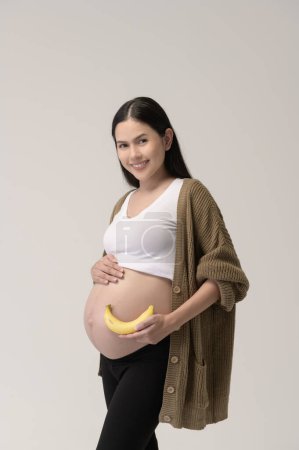Téléchargez les photos : Portrait de Belle femme enceinte tenant banane sur fond blanc studio, concept santé et maternité - en image libre de droit